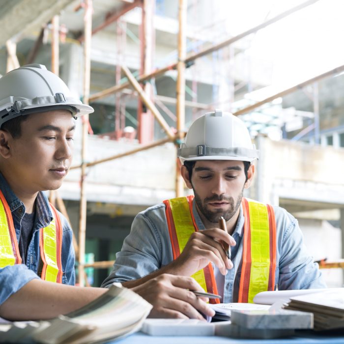 Young Asian engineers and caucasian engineer colleagues at site inspector and discuss together using notebook computer and paper blueprint at building construction site.home renovation ideas concept